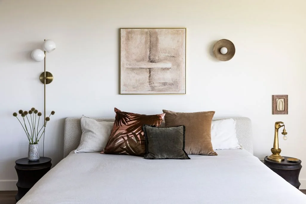 A pale upholstered bed with light bedding sits below one abstract picture beside dark round tables, small wall objects, warm-toned lamps and brown patterned cushions