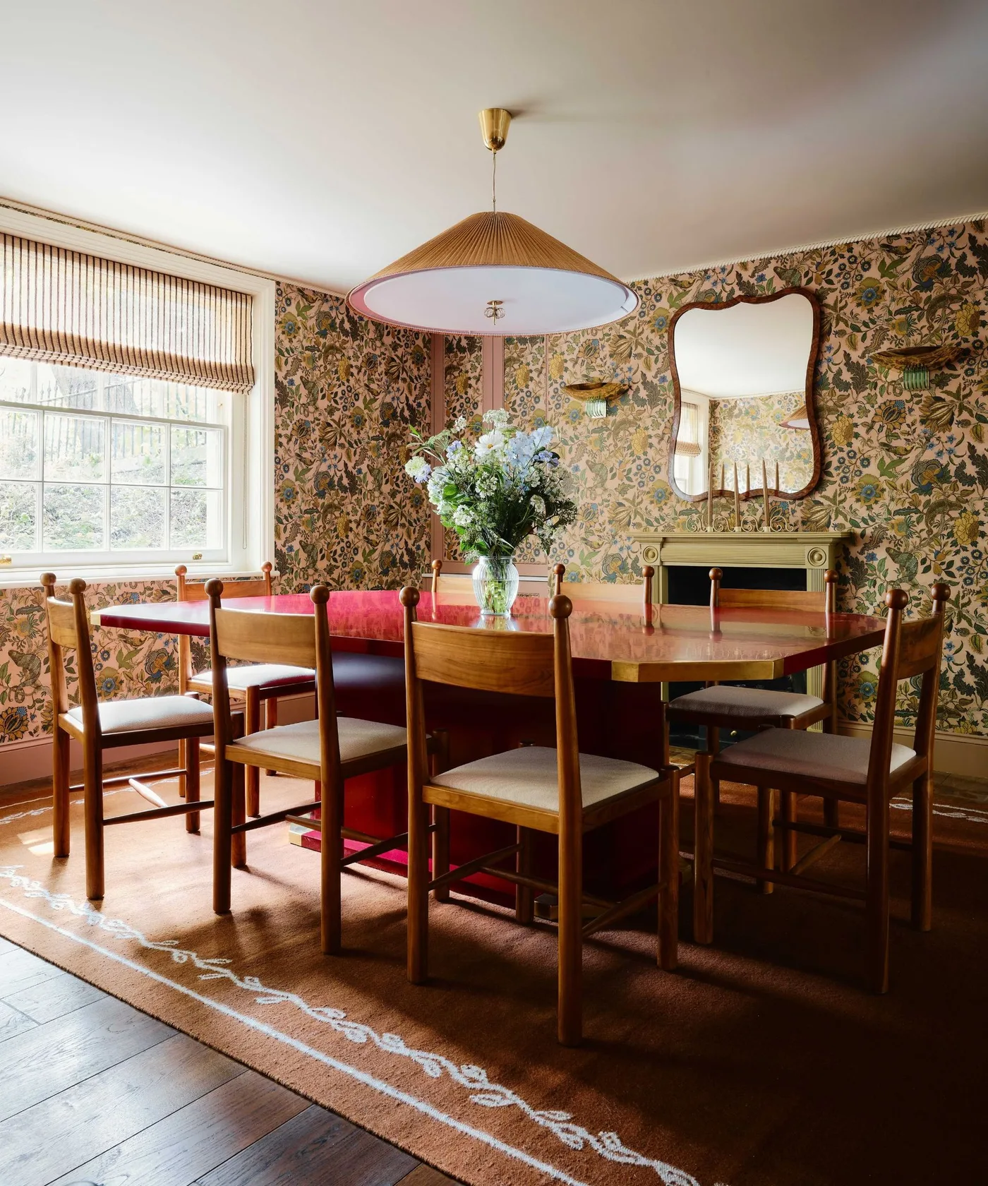 Country dining room with floral wallpaper, red table, wooden chairs with pale seats, broad pendant and a curved wall mirror.