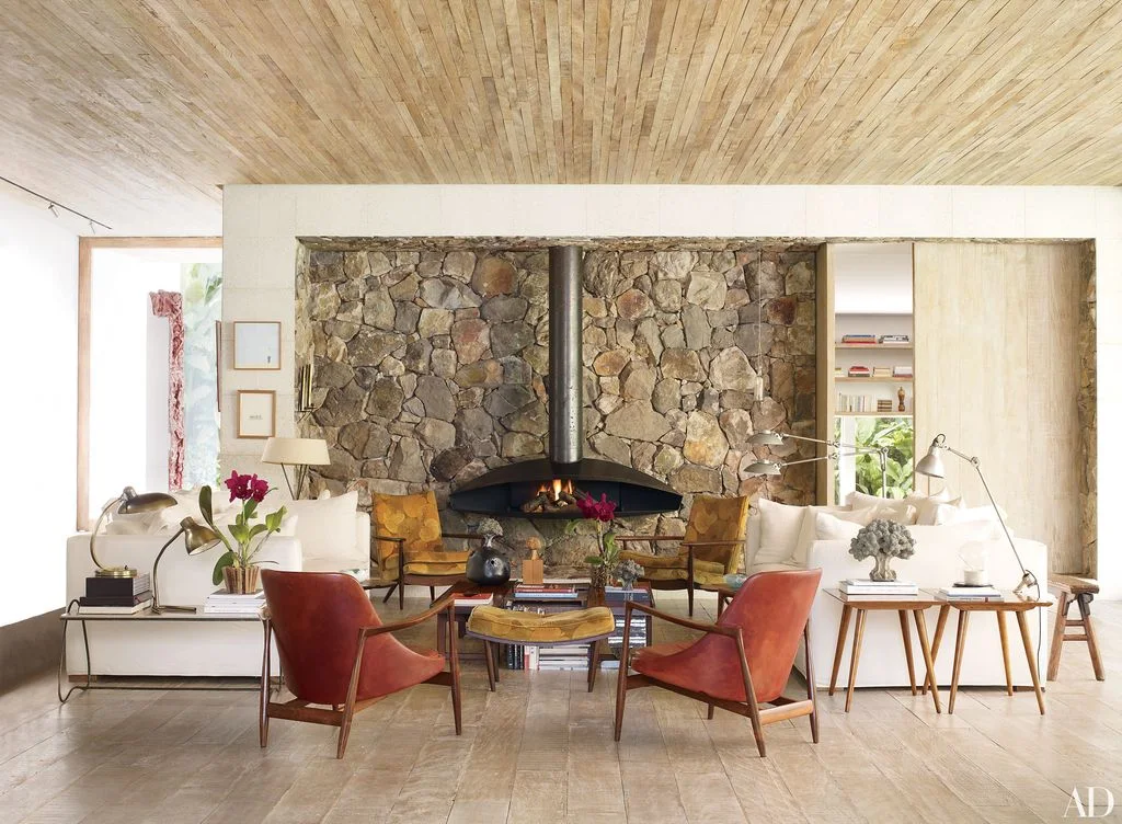 A dark sculptural fireplace hood and vertical flue stand in front of a broad rough stone wall beside pale sofas, red chairs and wood-framed seating