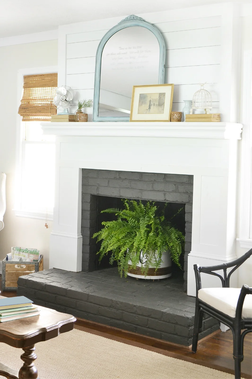 A white mantel surround stands against horizontal white boards above gray painted brick, with a pale blue arched mirror and a fern in the unlit opening.