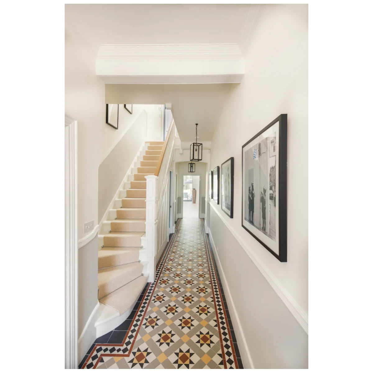 Long narrow entrance hall with geometric floor tiles, pale walls, framed pictures and a staircase along one side.