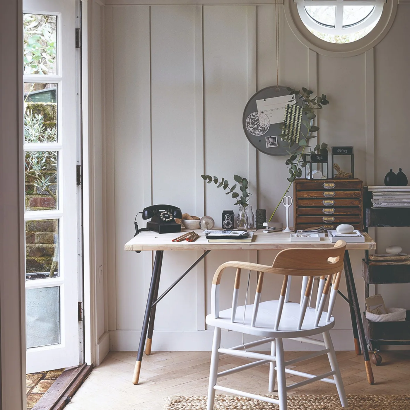 A pale desk with dark angled legs stands against white vertical boards, with a partly white-painted wood chair, shallow drawers and a round wall organizer beneath a circular window.