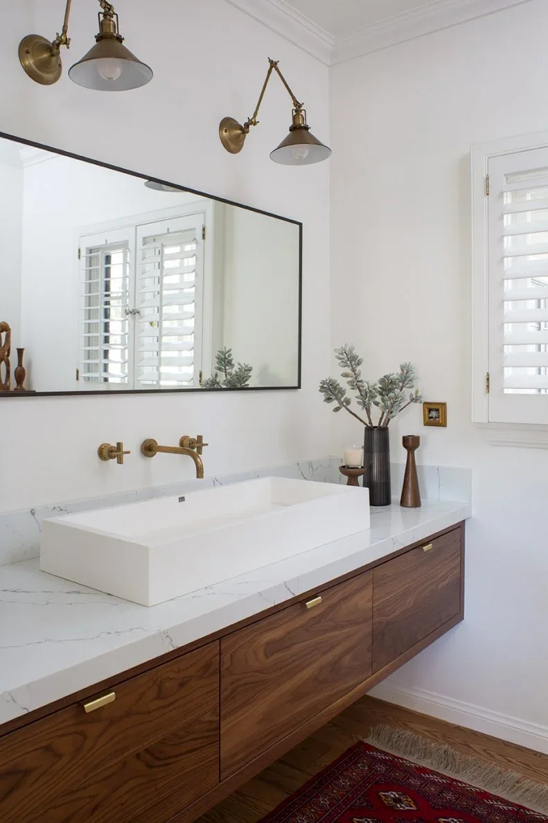 A wood-fronted floating bathroom vanity holds a wide white basin beneath a dark-edged mirror and two gold-colored wall lights beside a shuttered window