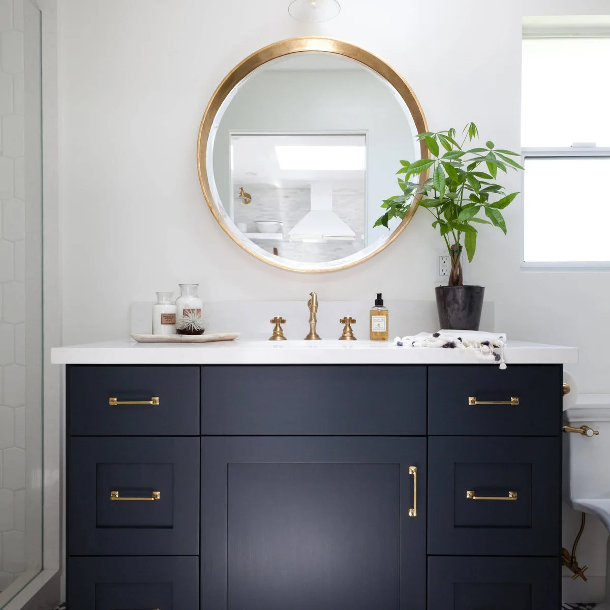 Dark vanity with a pale bottle tray on the left and a small potted tree beside a window on the right.