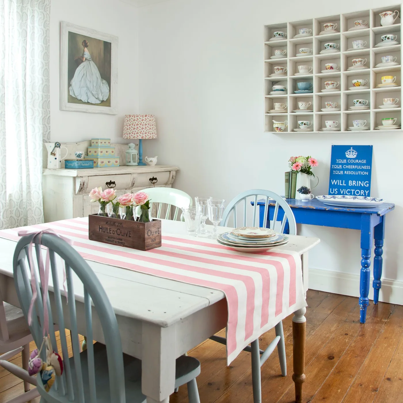 A white wall rack displays teacups above a small blue console beside a pale dining table with a pink striped cloth and pastel chairs.