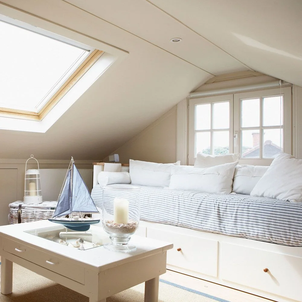 Striped daybed with drawers beneath a dormer window in a pale attic room with a coffee table.