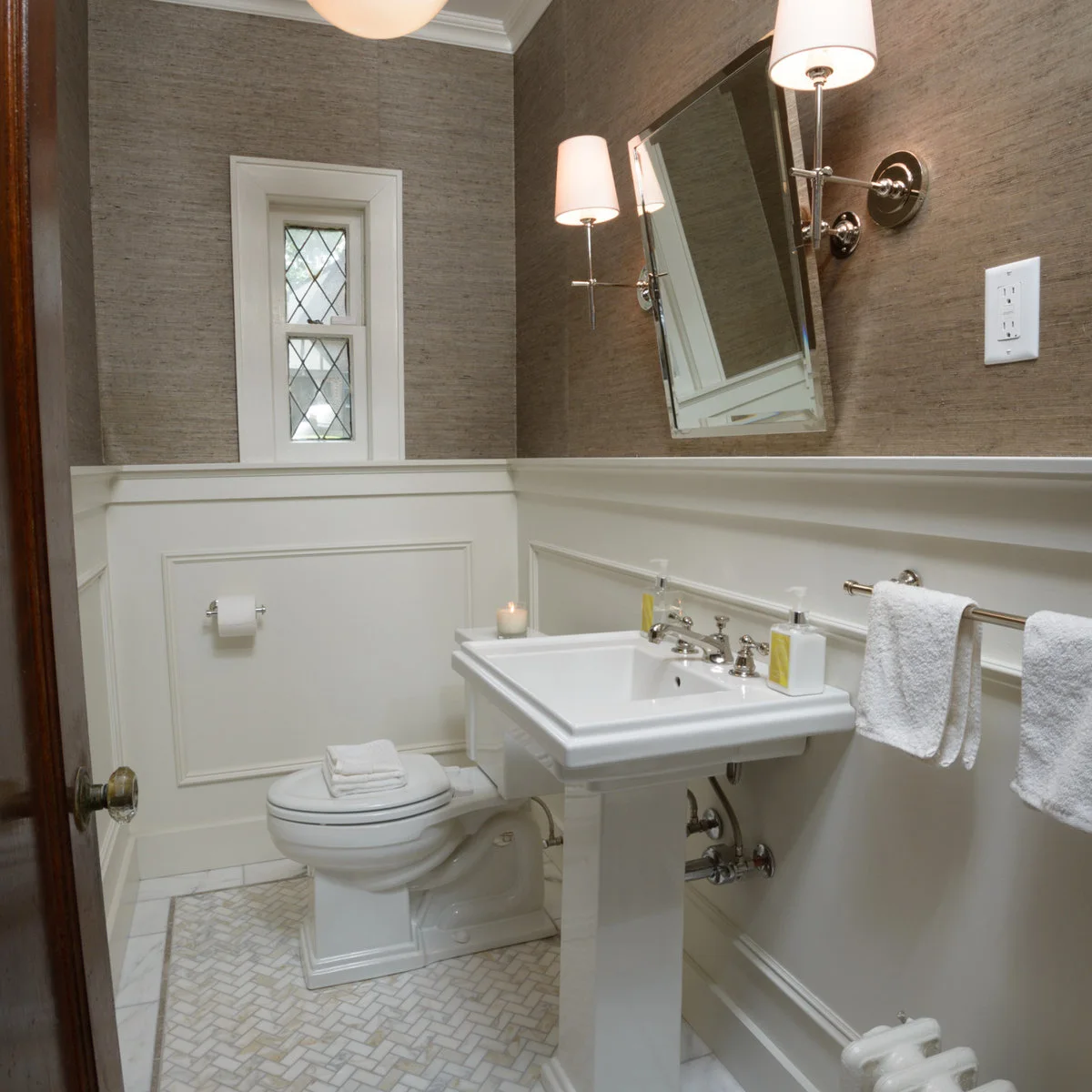 A white pedestal sink sits below a tilted rectangular mirror against white paneling and warm textured walls, with a long towel rail beside it.