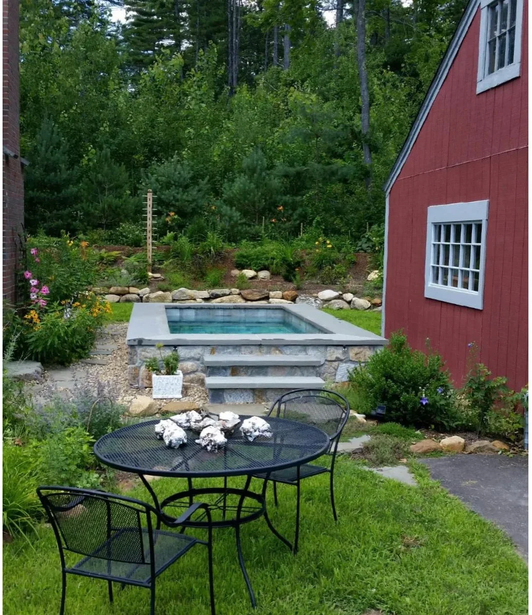 A raised stone-faced plunge pool with broad front steps sits beside a red outbuilding in a flower-filled courtyard.