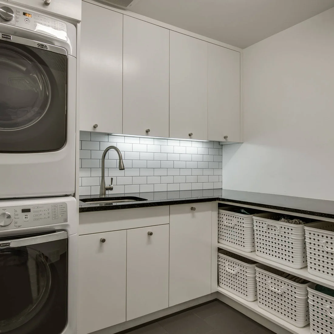 Stacked white machines and white cupboards sit beside open shelves of white baskets beneath a dark counter and white brick-pattern backsplash.