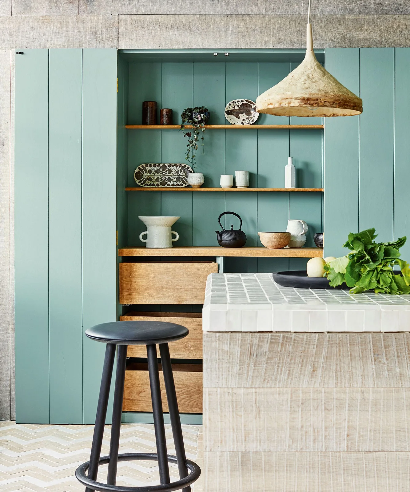 A turquoise kitchen recess contains wooden shelves, pale ceramics and drawers beside a tiled island
