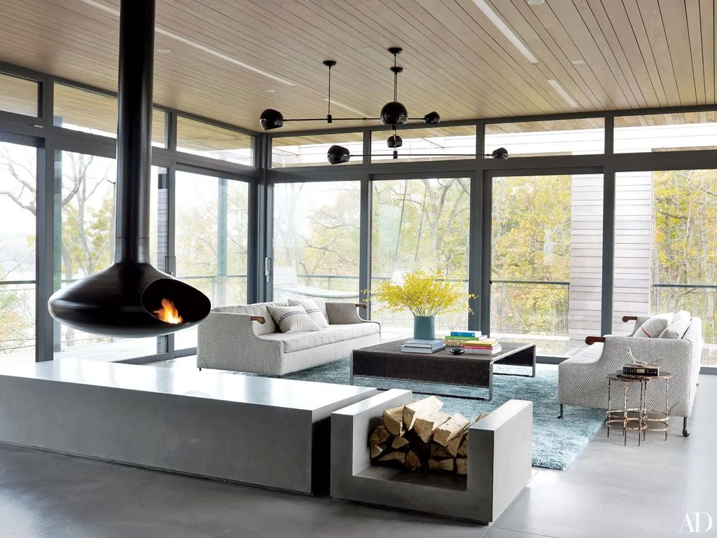 A rounded dark suspended fireplace hangs from a long flue beside pale sofas, broad black-framed windows, a blue rug and a low pale wood-storage divider