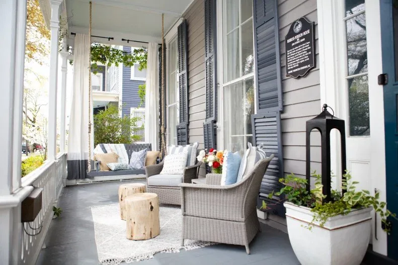 Gray porch with pale woven armchairs, stump-shaped tables, white rug and swing at far end