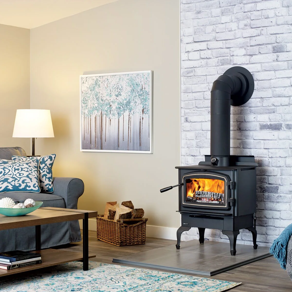 A black wood stove stands on a gray rectangular hearth against a pale brick wall beside a blue-gray sofa and patterned rug.