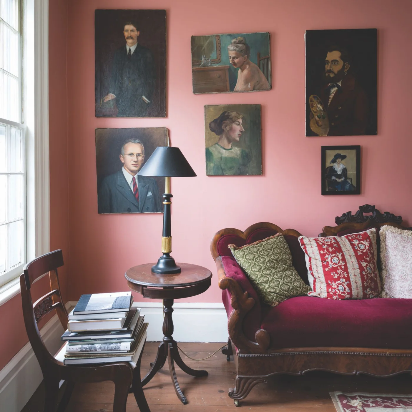 A carved wooden sofa with deep pink upholstery sits below framed portraits beside a round wooden table