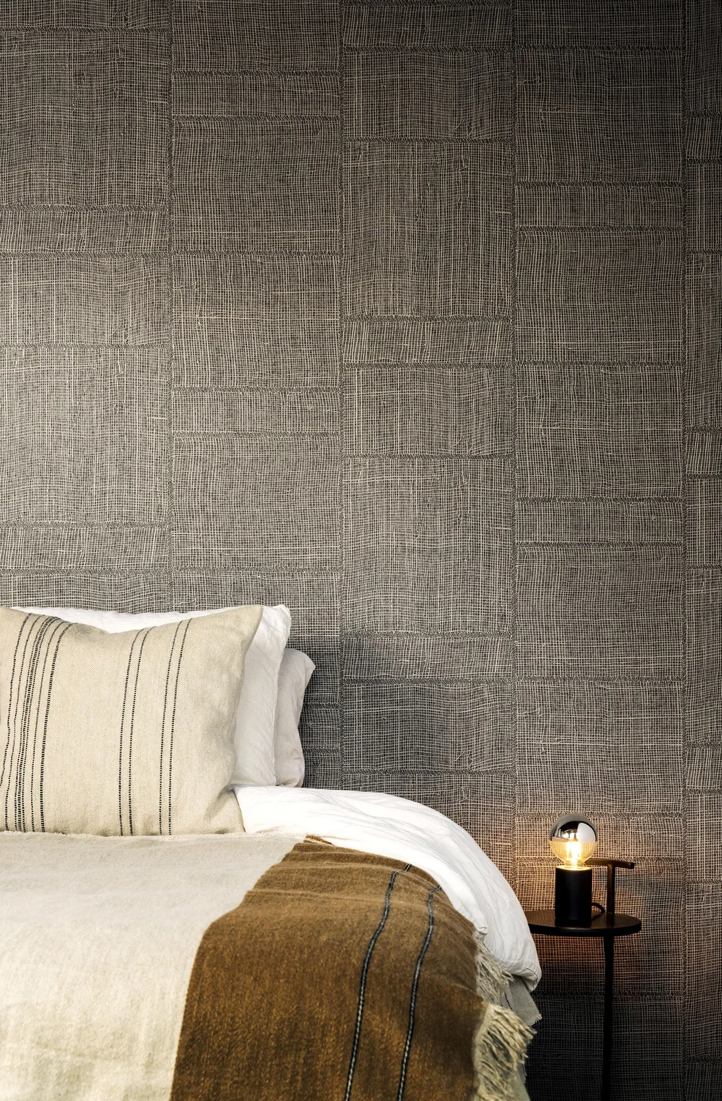 A dark gray textured wallpaper in irregular rectangular sections rises behind pale bedding, an ochre throw and a small dark bedside table with a glowing bulb lamp.