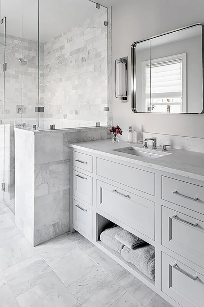A pale bathroom vanity with drawers and an open towel compartment sits beside a glazed shower, small pale wall tiles and a dark-edged mirror