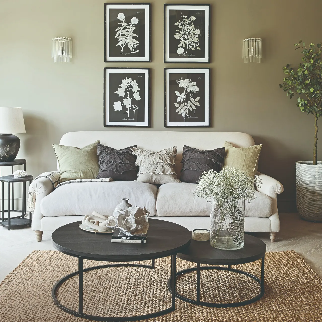 A pale sofa sits beneath framed botanical pictures beside a dark round table