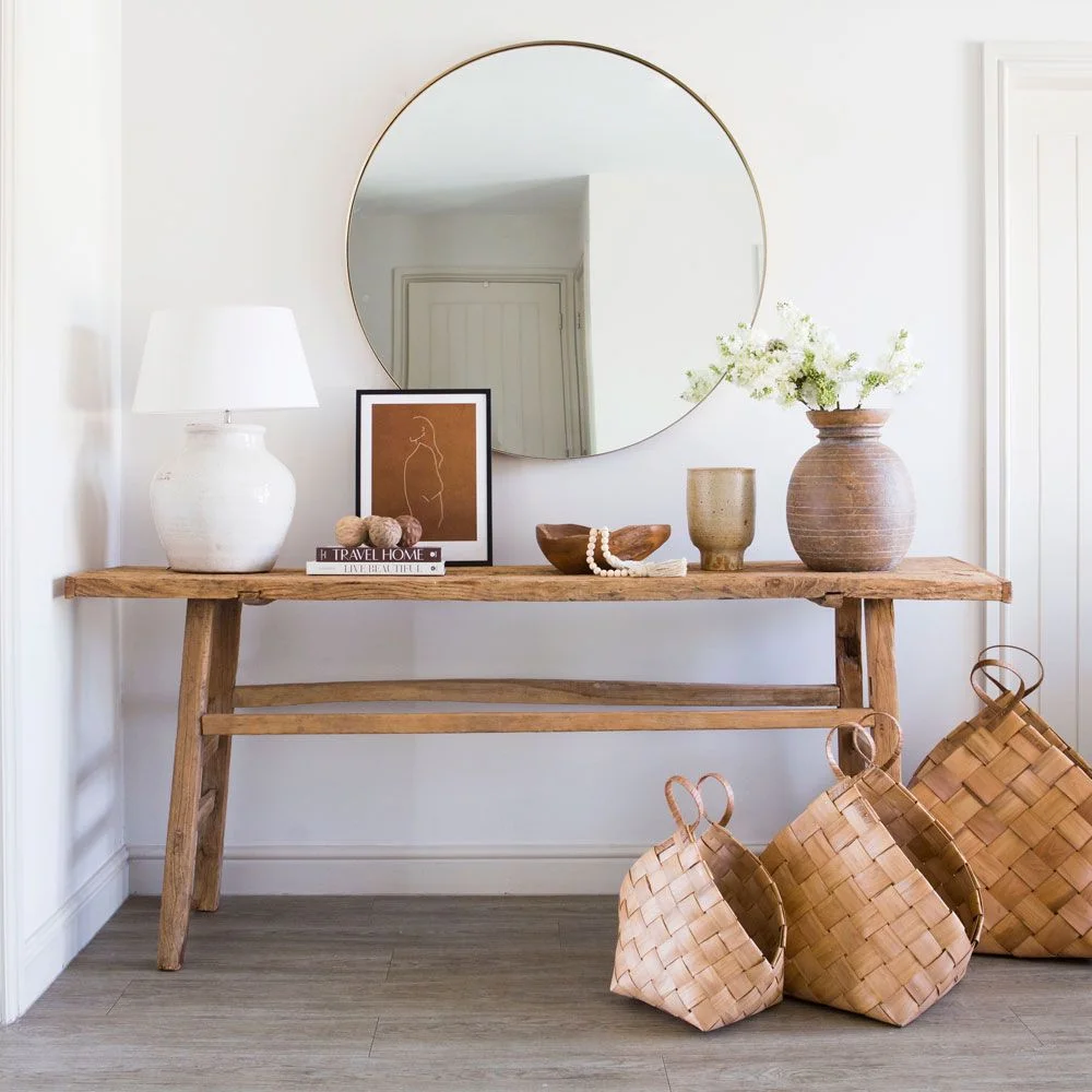 A rough-edged wood console stands beneath a round mirror beside pale pottery, a shaded lamp and woven floor baskets.