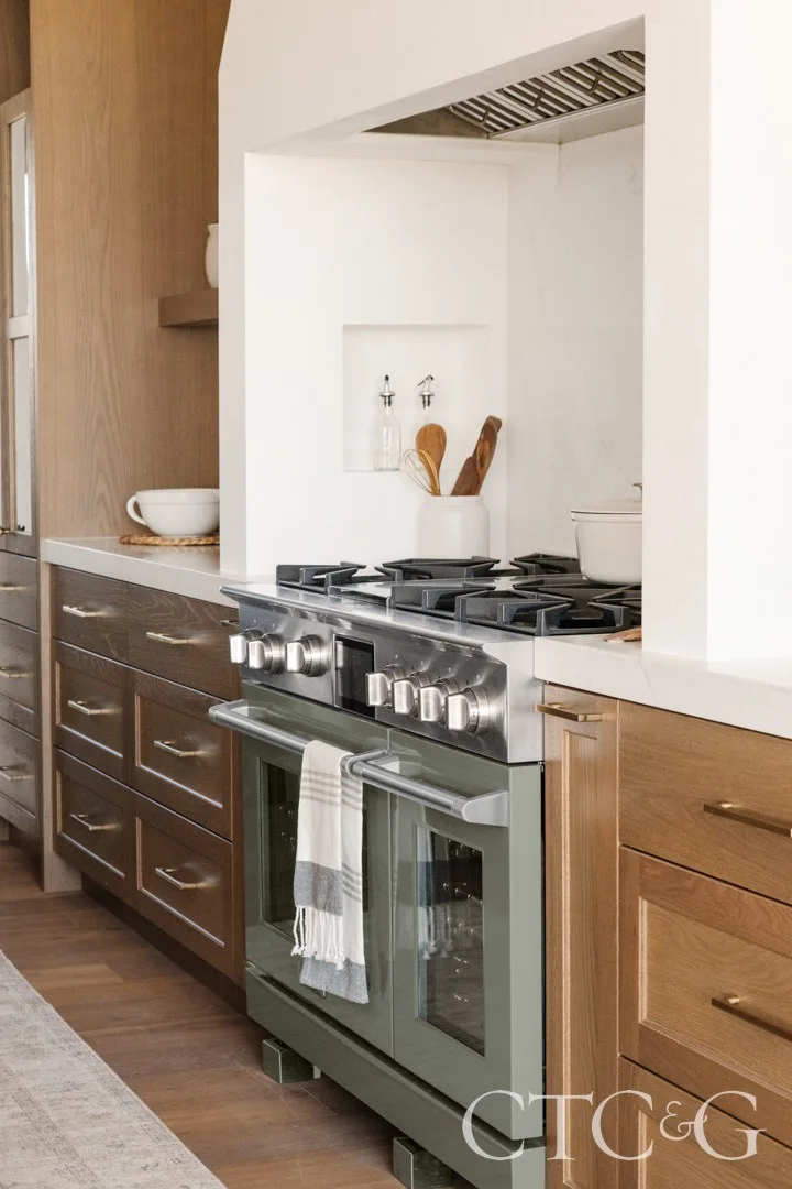 White oak drawers with brass-colored pulls sit beside a muted green range within a white hood recess and pale countertop.