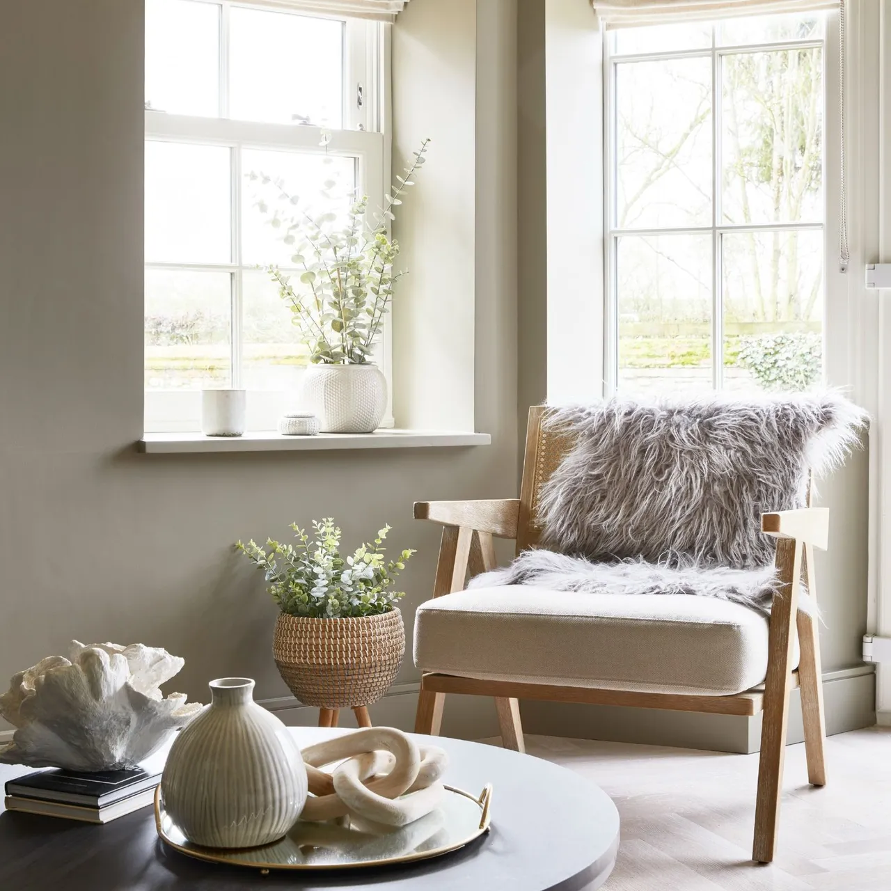 A pale wood armchair with a textured gray throw stands between bright windows, beside a raised woven planter and a dark round table with pale objects.