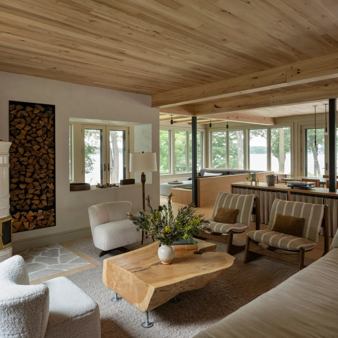 An irregular wood slab coffee table stands among pale chairs beneath a broad wood ceiling in a window-lined living room.