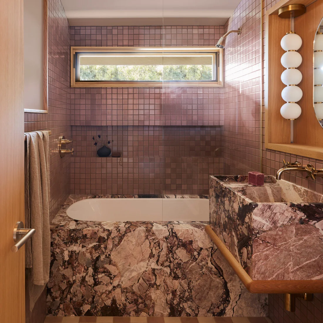Small rose-colored square tiles line a bath beneath a horizontal window beside a heavily veined pink counter