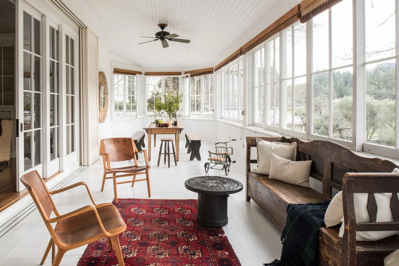 A darker sofa sits beside timber-framed chairs on a red patterned rug in a bright sunroom.