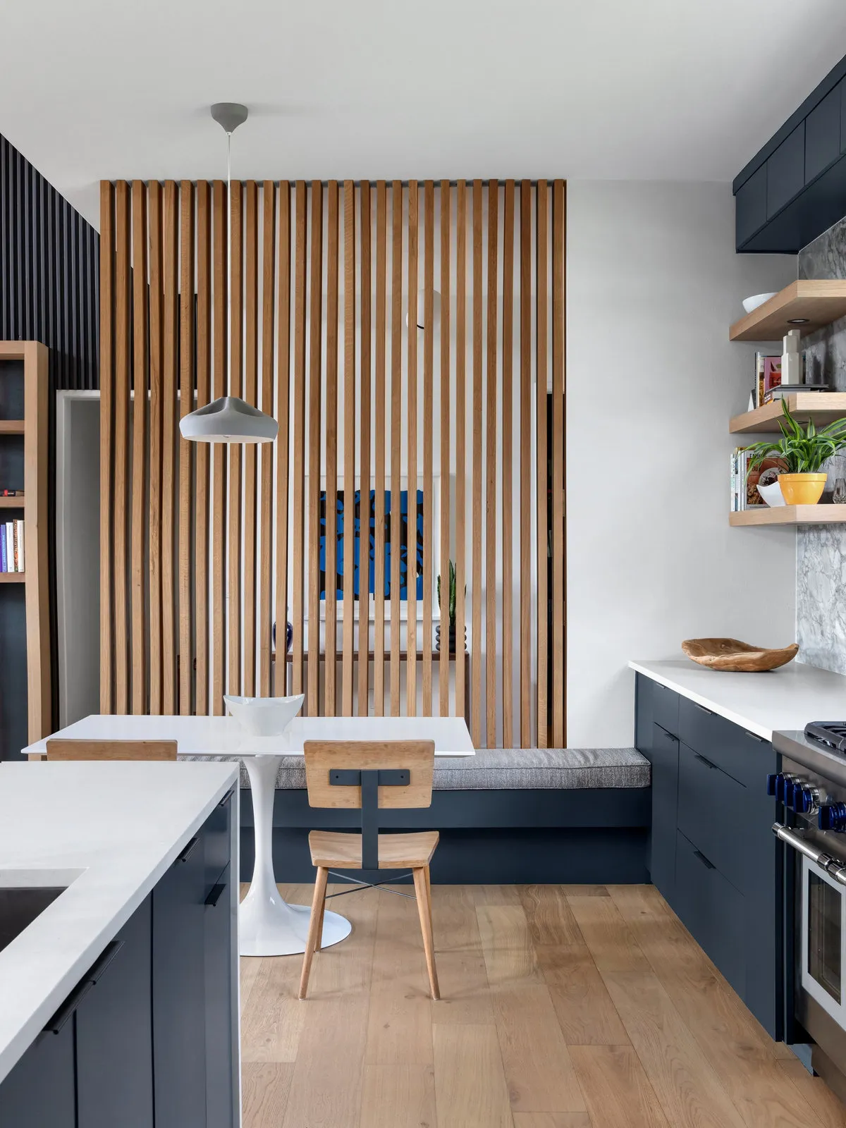 Blue cupboards and pale counters sit beside a dining nook backed by an open timber slat divider.