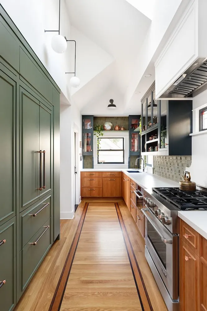 Warm cherry lower cabinets run opposite a tall green storage wall in a narrow kitchen