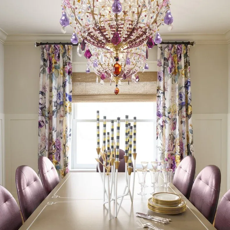 Dining room with a multicolored hanging chandelier, purple upholstered chairs, floral curtains and a reflective rectangular table