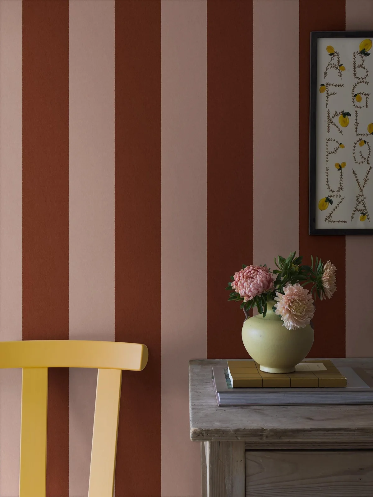 Broad rust and pale pink wallpaper stripes stand behind a yellow wood chair, weathered gray table, framed botanical alphabet and pale green flower vase.