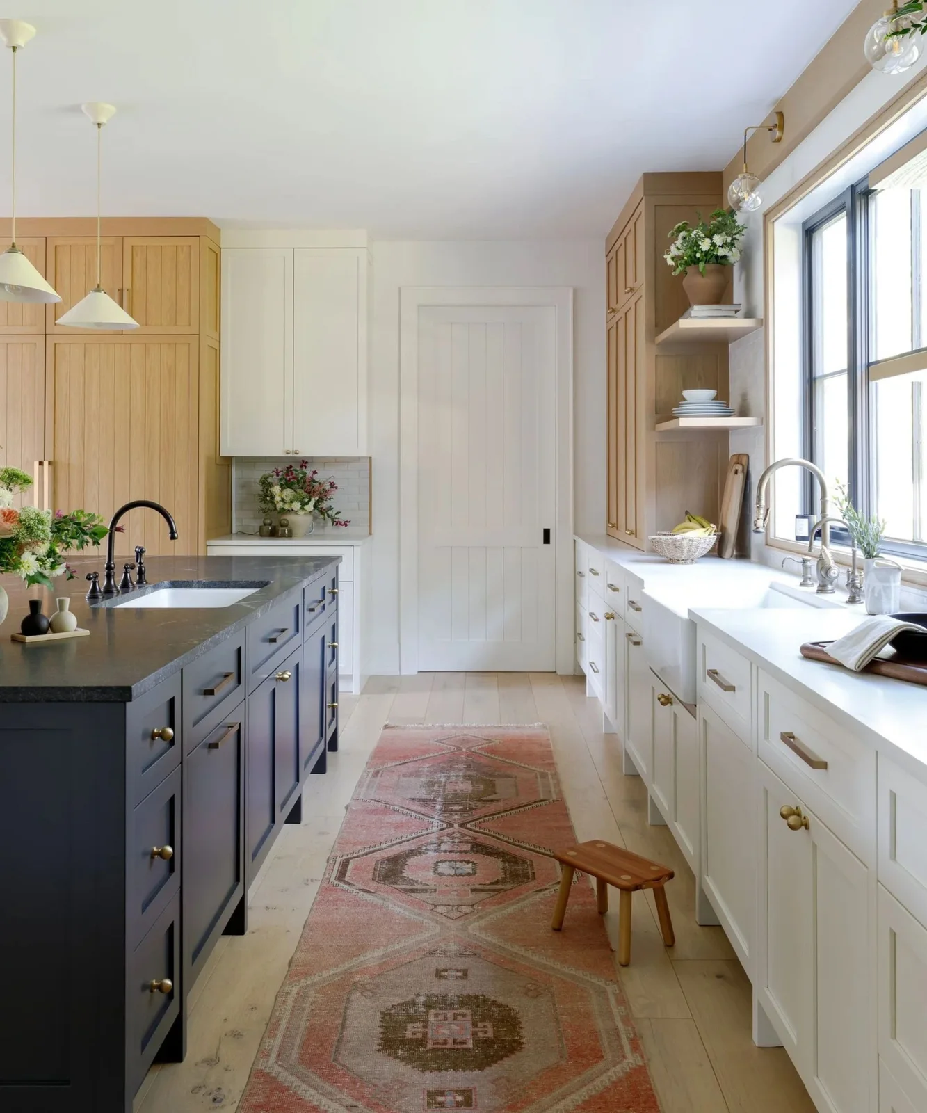 Natural oak tall and upper cabinets mix with white sink cabinets and a navy island in a bright kitchen with a patterned runner.