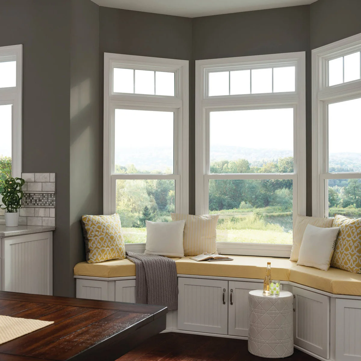 Yellow cushions on a white storage seat beneath three tall windows, beside a small round side table.