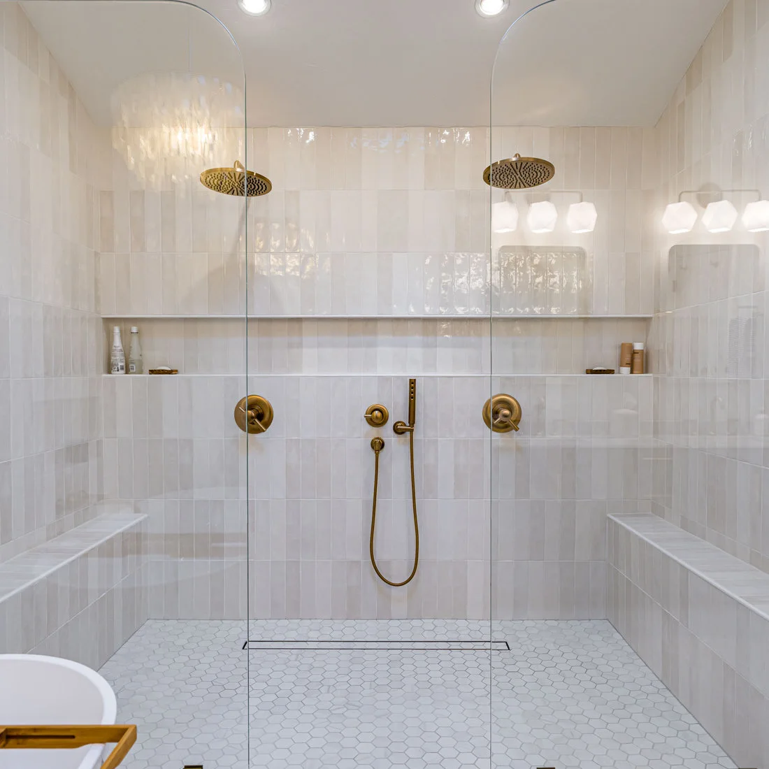 A wide glass shower has pale vertical rectangular tiles, two shower fittings and a long niche above a hexagonal floor