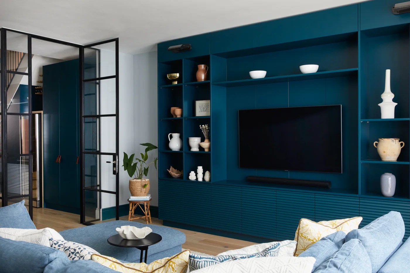 Deep blue fitted media storage combines ridged lower cupboards, open display shelves and a television beside black-framed doors.