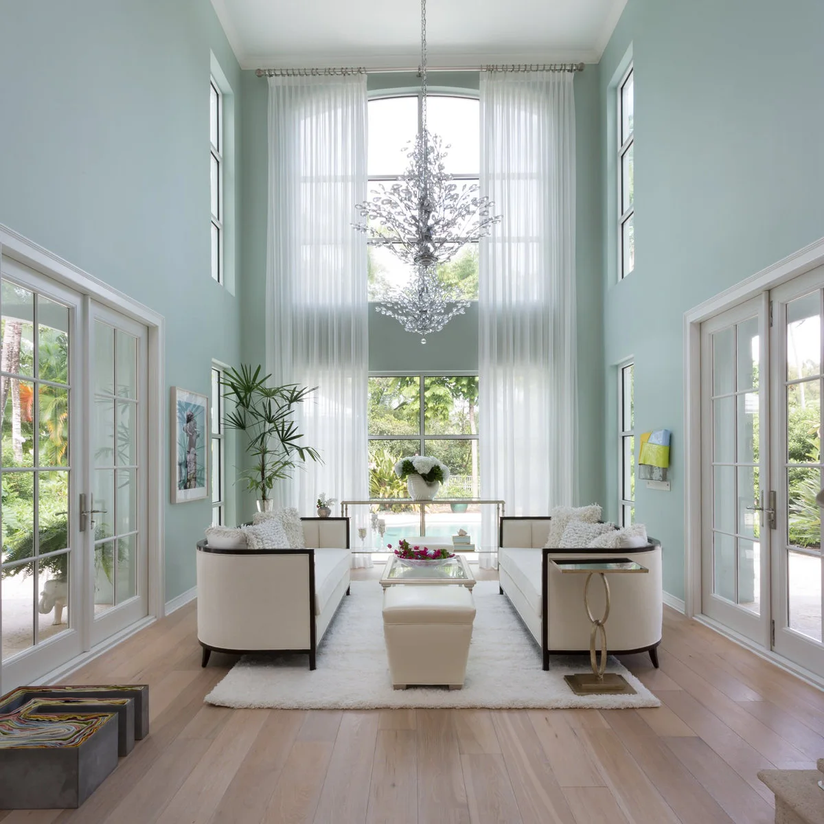 A narrow tall living room with pale blue-green walls, long white curtains, facing cream sofas and a cascading chandelier.
