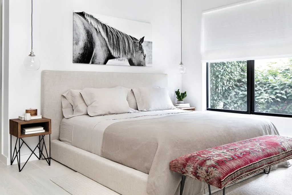 A pale upholstered bed with light bedding stands beneath a horse picture beside small wood tables and slim hanging lights with a pink patterned bench at the foot
