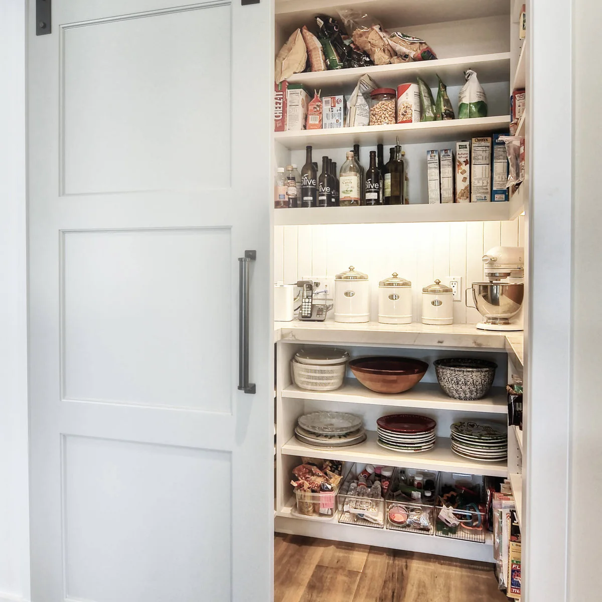 A pale sliding door opens onto food shelves, a lit counter with canisters and a mixer, and lower shelves of bowls and plates.