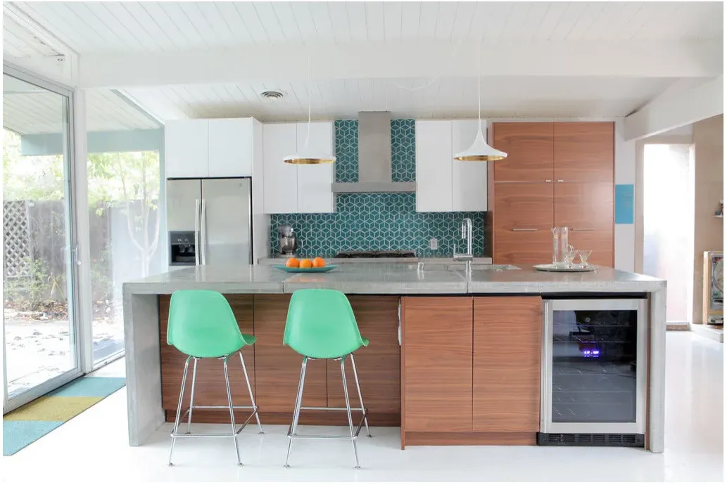 Two green shell-shaped stools at a wood-fronted island beneath white pendants, with geometric blue-green backsplash tile.