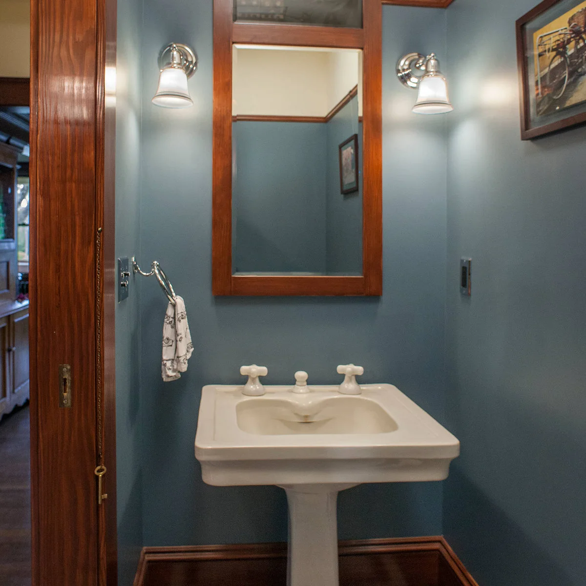 A small white pedestal sink stands beneath a wood-framed mirror in a narrow blue room with matching wood door trim.