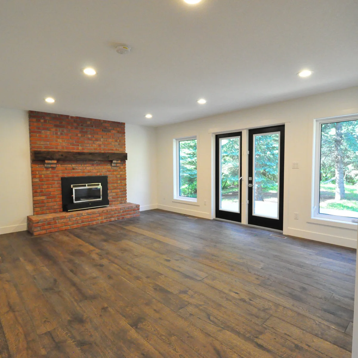 A retained red brick fireplace with a dark wood mantel stands beside pale walls, dark glazed doors and broad gray-brown floorboards.