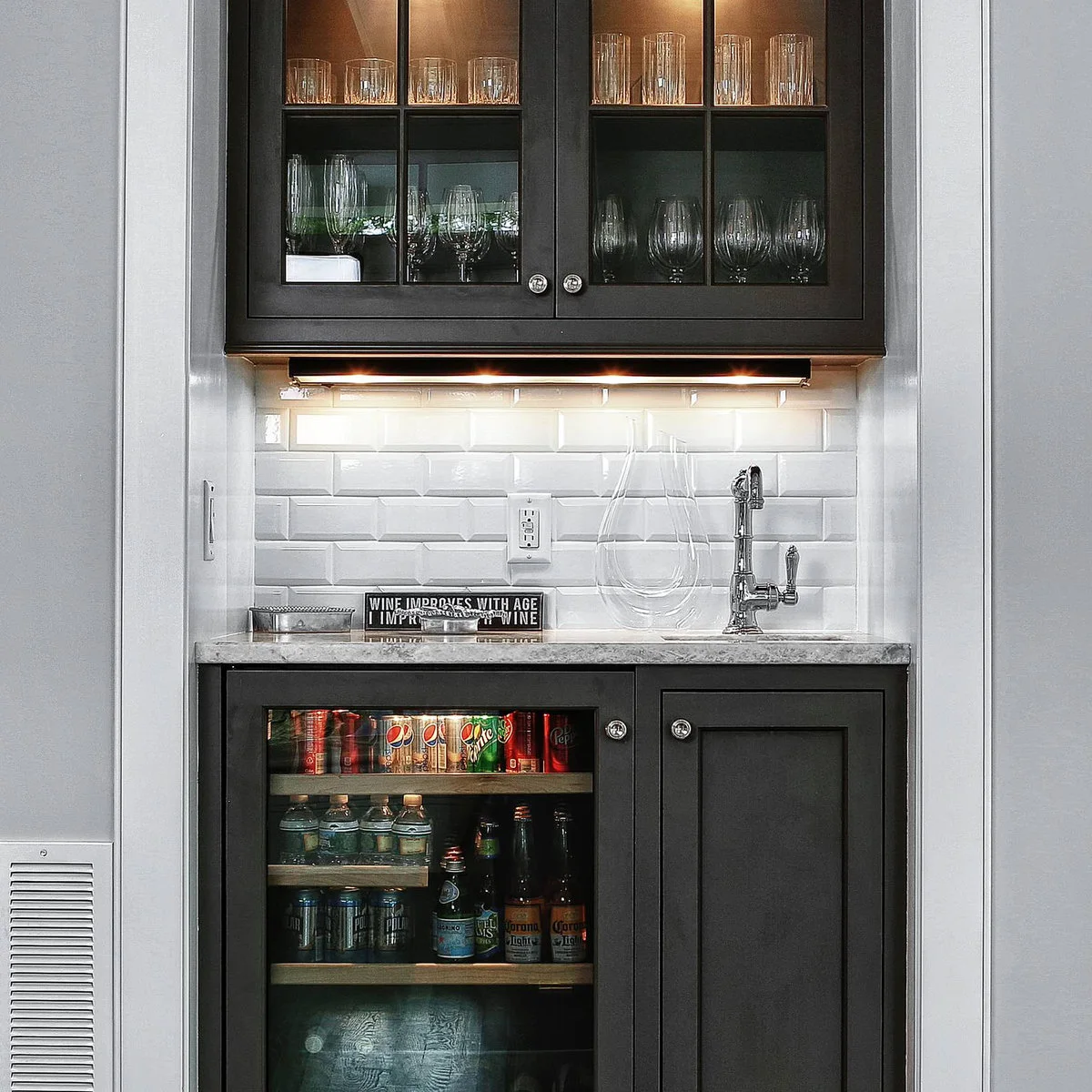 Narrow black mini bar with glass-front upper cabinets, white beveled tile, small faucet and beverage refrigerator