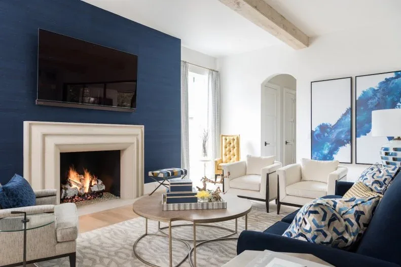 Living room with deep blue fireplace wall, dark blue chair, pale armchairs, paired blue abstract pictures, round table and pale wooden ceiling beam.