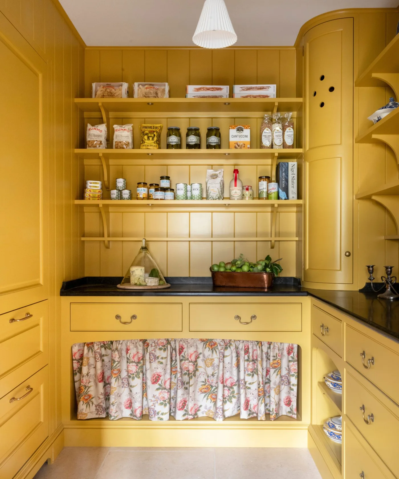 Yellow country pantry with open shelves, dark counter, lower drawers and floral cabinet skirt