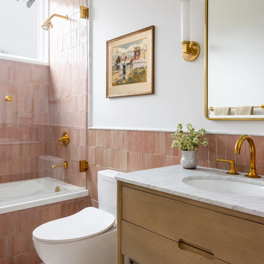 Pale pink rectangular tiles surround a bath and sink below white walls beside a wooden vanity and gold-colored fittings