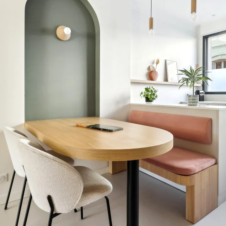 A pale wood oval-ended table meets a pink cushioned bench beside a sage-green arched recess and two pale chairs.