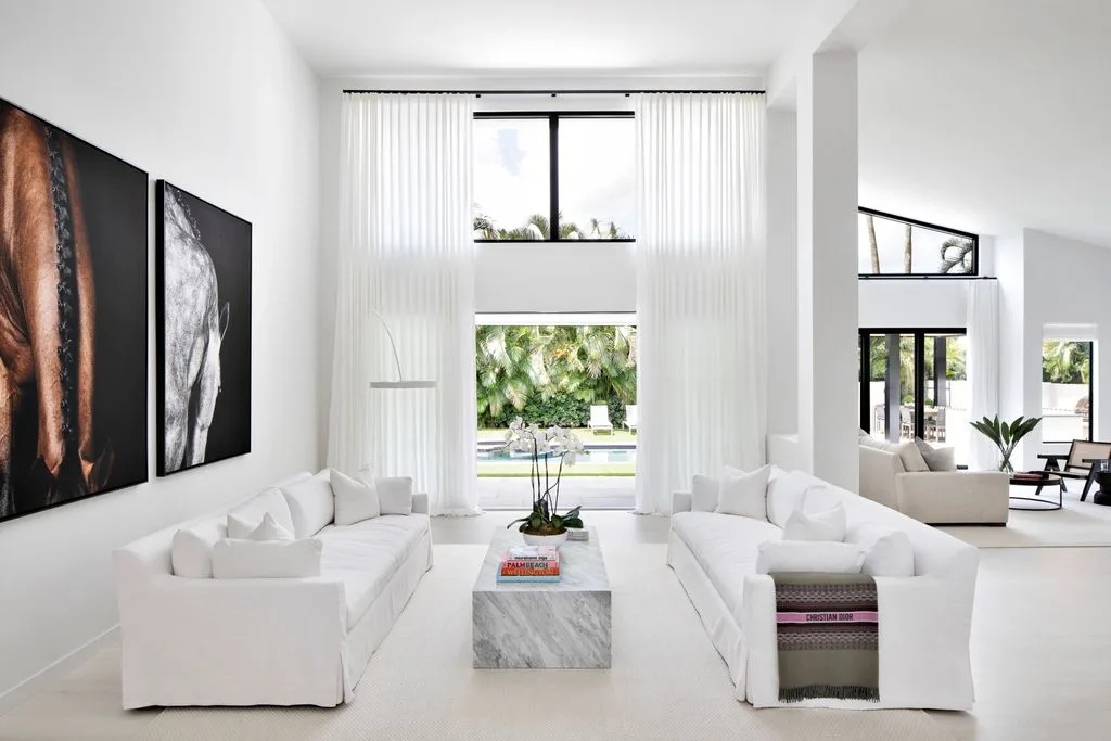 Double-height pale living room with two facing white sofas, a rectangular coffee table, large wall photographs and tall curtains.