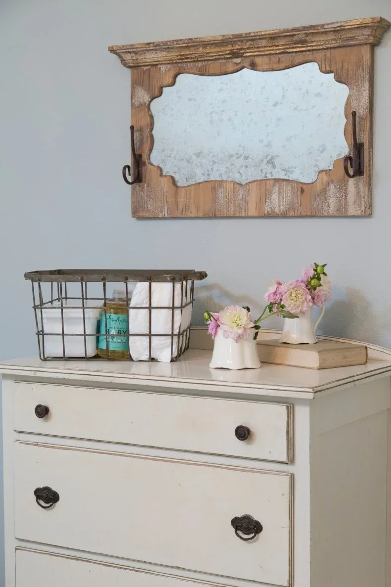 Painted nursery dresser beneath rustic wood mirror, with wire basket, folded cloths and small flower jugs