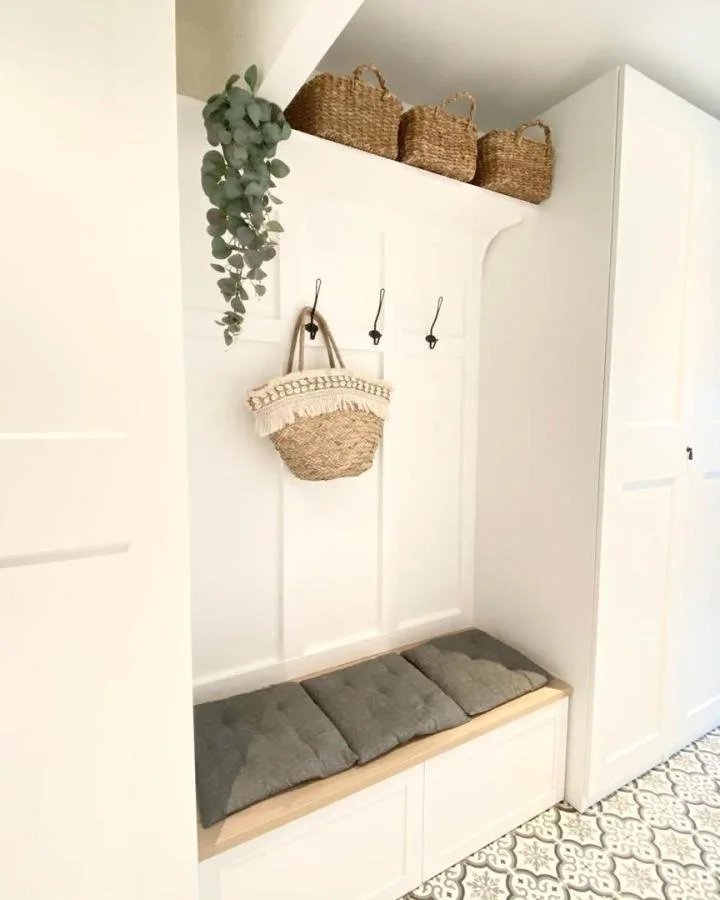 Compact white storage bench with two gray cushions beneath three black hooks and a high shelf of woven baskets.