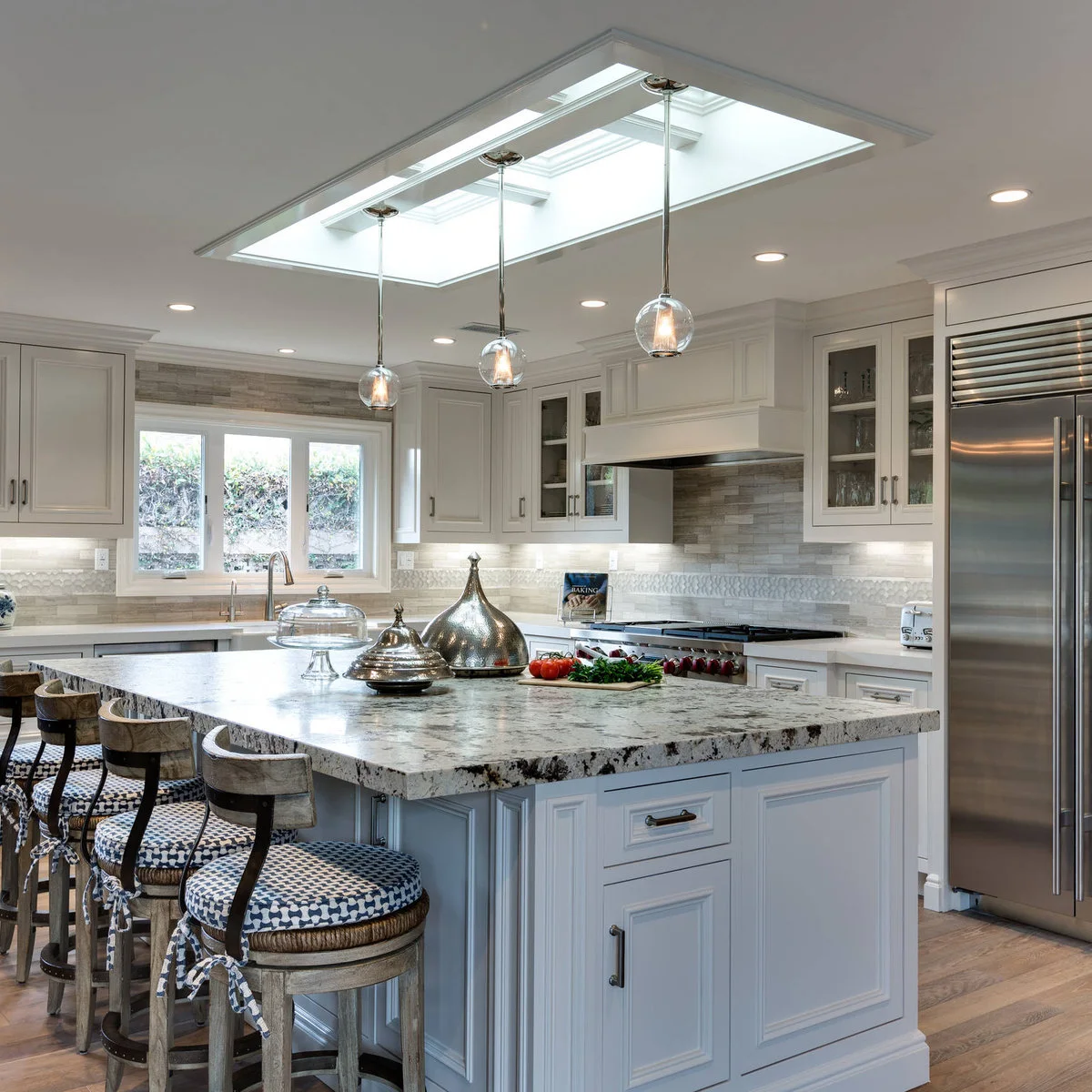 Pale kitchen island with a patterned top beneath a long skylight and glass pendants
